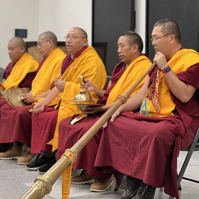 Five monks sitting in a line of chairs dressed in dark red and yellow robes. One is holding a long musical instrument, the others have smaller instruments in their hands..